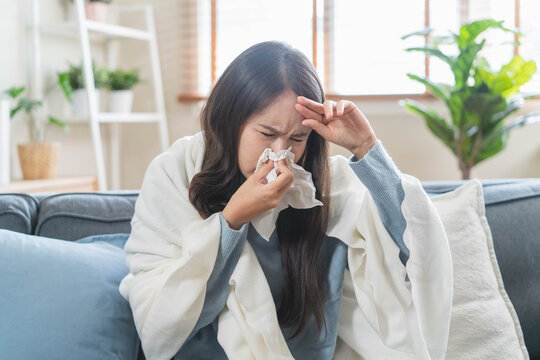 Sick Asian woman blowing her nose with tissue on sofa, wrapped in blanket, suffering from high fever, seasonal flu, influenza virus infection, and cold symptoms at home