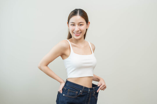 Happy slim Asian woman showing weight loss success by pulling loose big jeans on waist, smiling healthy young female after fitness workout and diet motivation concept.