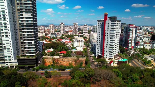 High-angle aerial drone shot flying forward and tracking right parallel to an avenue, showing a vacant lot framed by residential skyscrapers and park trees.