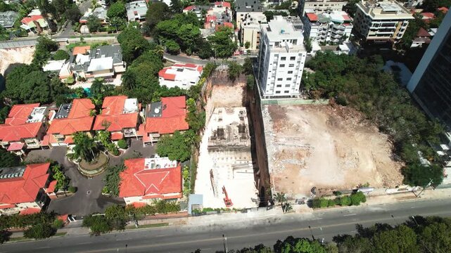 High-angle aerial drone shot advancing and tilting down to top-down zenith view over a building excavation site with drilling machinery next to avenue and park trees