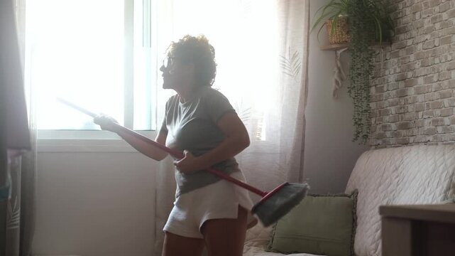 Joyful woman having fun while doing housework, singing and dancing with a broom like a guitar in her living room. Overjoyed female person enjoying domestic chores and cleanliness