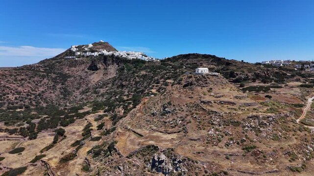Klima lies below dry volcanic hills marked by eroded pyroclastic slopes and exposed rock, with white Cycladic buildings on the ridges, Milos, Greece, drone forward flyover.