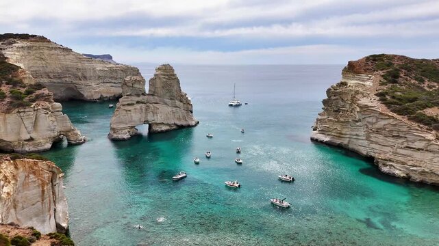 Kleftiko sea caves and volcanic rock formations frame a turquoise cove filled with anchored motorboats and sailboats on the Aegean Sea, Milos, Greece, drone pullback.