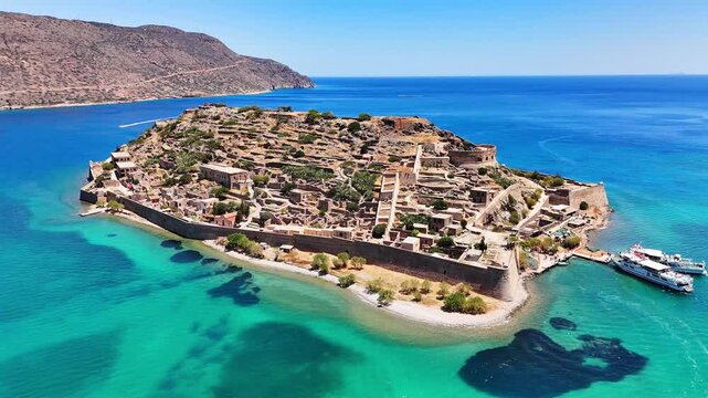 Kalydon Island and Spinalonga fortress show Venetian walls, stone ruins and moored motorboats along a turquoise shoreline in Mirabello Bay, Cretan Sea, Lasithi, Greece, drone orbit establishing shot.