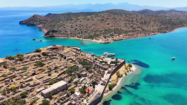 Kalydon Island and Spinalonga fortress rise above turquoise Mirabello Bay, with Kolokytha Island and the Cretan mountains beyond, Lasithi, Crete, Greece, drone orbit parallax.