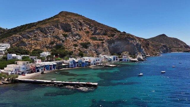 Klima fishing village lines the turquoise coast with colorful syrmata boathouses beneath rugged volcanic cliffs, facing the Aegean Sea on Milos, Greece, lateral drone slide toward the coast.