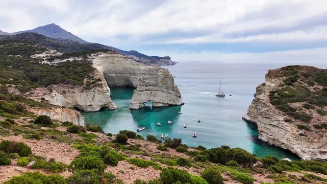 Kleftiko features white volcanic cliffs, sea caves and natural arches surrounding a turquoise cove with motorboats and a sailboat on the Aegean Sea, Milos, Greece, Drone Forward Flyover.