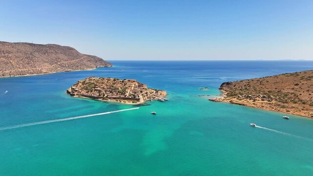Kalydon Island and Spinalonga fortress sit between rugged Cretan coastlines, with motorboats leaving wakes across turquoise Mirabello Bay in the Cretan Sea, Lasithi, Greece, drone slide.