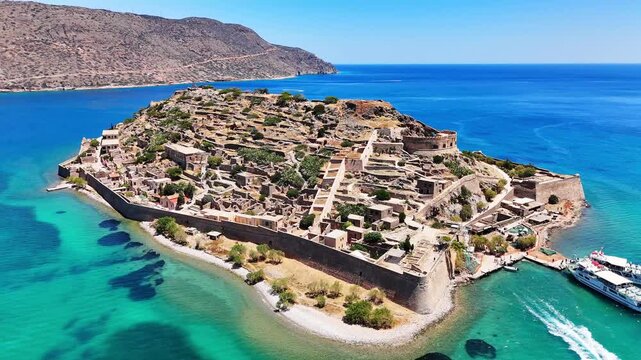 Kalydon Island and Spinalonga fortress display Venetian walls, stone ruins and a harbor with moored motorboats along the turquoise coast of Mirabello Bay, Lasithi, Greece, drone push in flyover.