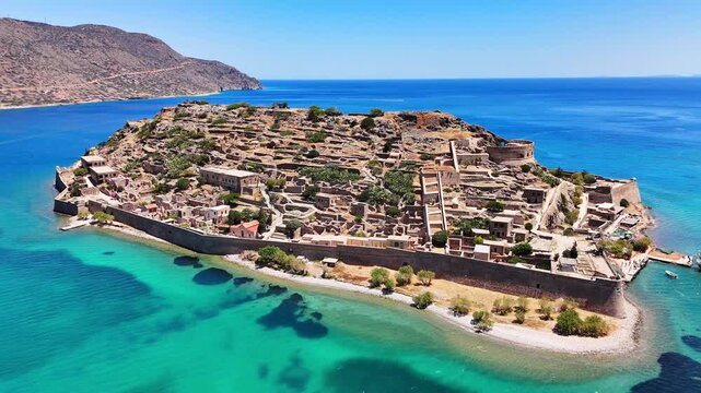 Spinalonga Fortress and Venetian ruins cover Kalydon Island, surrounded by turquoise waters of Elounda Bay with rugged Crete mountains beyond, Greece, drone pullback.