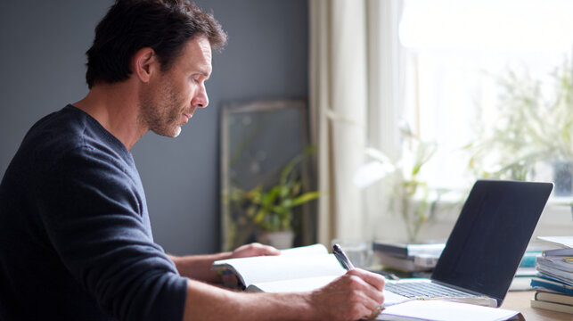 Premium lifestyle photograph of an adult man learning a new skill through an online course at a home desk, laptop open but screen facing away from camera, notebook and pen beside it