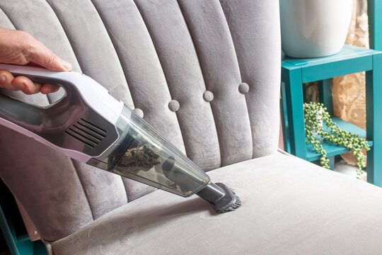 Hoovering vacuuming the upholstery on a grey chair with a handheld cordless vacuum cleaner. Household cleaning chores concept.