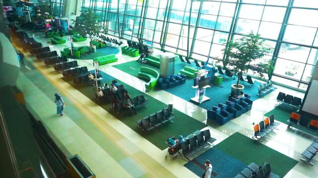High angle wide view of modern spacious airport terminal lounge seating area with green carpeted waiting zones glass facade windows and passengers waiting for flight boarding inside departure hall