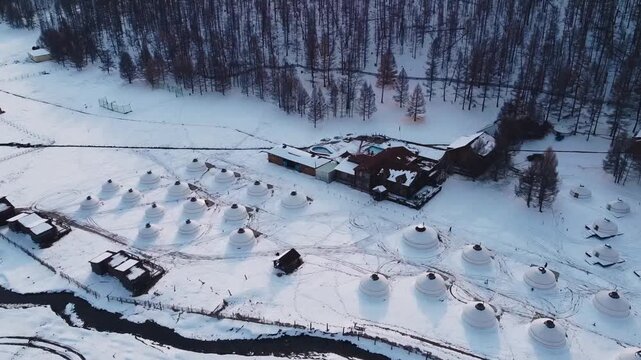 Aerial view of a traditional winter settlement with wooden buildings and snow-covered igloos surrounded by dense coniferous forest in Mongolia