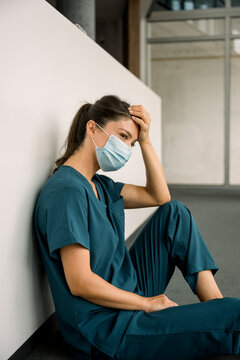 Exhausted nurse in scrubs and face mask sitting on hospital floor