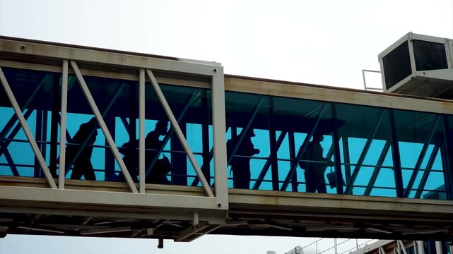 Air Passengers Walking Along Aerobridge Jet Bridge Passage Way Boarding Aircraft Glass Window Airport Passenger Bridge Silhouetted Passengers In Terminal Transit Zone International Travel Concept