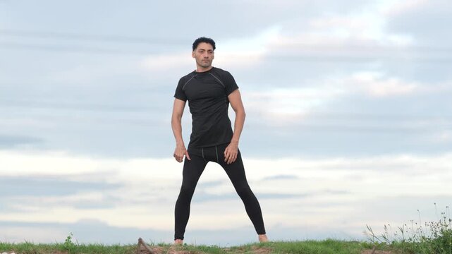 Man stretching exercise fitness outdoor grass hill under cloudy sky, athletic black sportswear side lunge warmup, rear view hands on lower back, yoga flexibility routine, calm focus soft evening light