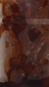 4K Vertical video melting ice cubes mixed in dark brown iced coffee inside glass, abstract liquid textures and reflections for cafe beverage menu concept.