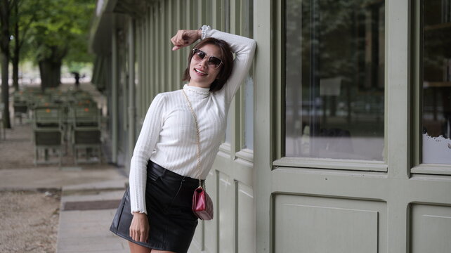 A stylish Asian woman wearing a white ribbed turtleneck sweater, black leather skirt, and sunglasses poses standing against a sage green building exterior in a park setting