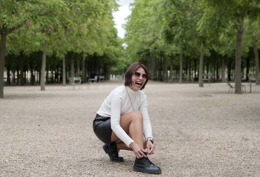 A cheerful Asian woman wearing a white turtleneck sweater, black leather skirt, and boots laughs while crouching down to adjust her shoelace along a gravel path in a park lined with green trees