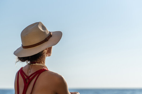 Rear view of young traveler in straw hat looking out at the calm ocean under clear sky