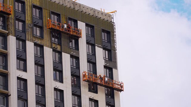 Construction workers installing facade on modern high-rise apartment building using suspended platforms, exterior cladding and insulation system, urban residential development concept