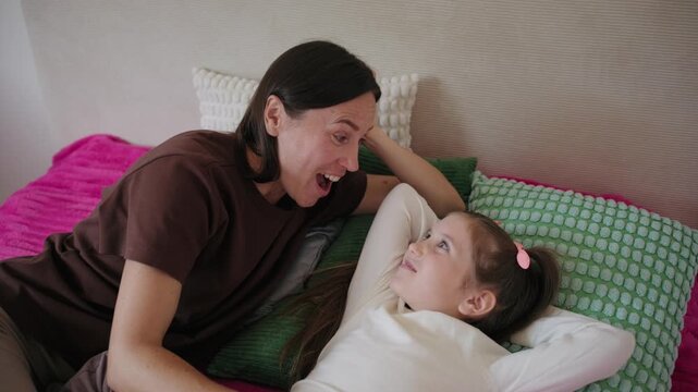 Caring mother talking and laughing with her little daughter while lying together on a bed at home.