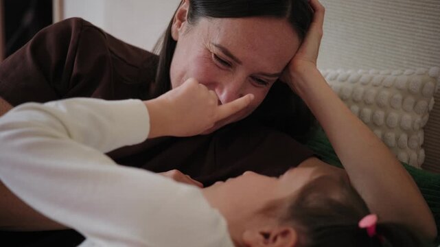 A loving mother and little daughter lie in bed together, talking, smiling, and playfully touching noses in a cozy bedroom.