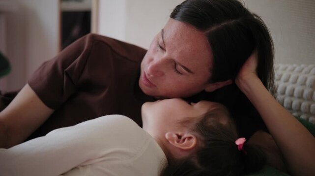 Loving mother talking with her little daughter while lying down together on a bed at home.