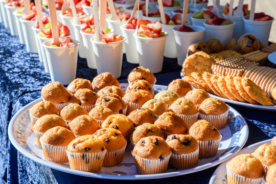 Cupcakes, cookies, and individual fruit salad cups arranged on a table during an outdoor picnic. The assortment of sweet treats and fresh fruit is prepared for casual social gathering in a relaxed out