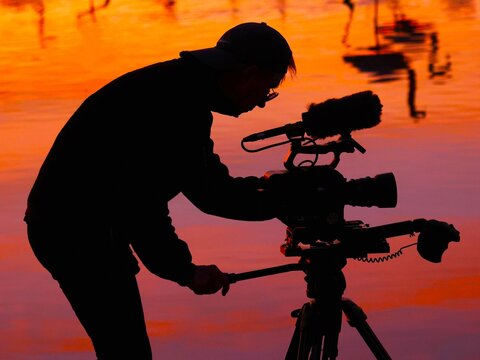 Vue de profil de la silhouette d'un homme r&eacute;glant le tr&eacute;pied de son appareil photo pour une prise de vue. Personnage en noir sur un fond orang&eacute;.