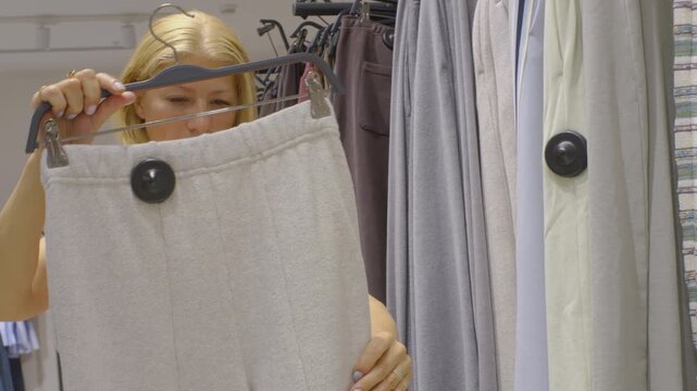 Side view of a female customer checking price tags and materials of neutral-toned pants during a shopping session in a clothing department. She holds the hanger firmly with one hand while adjusting th