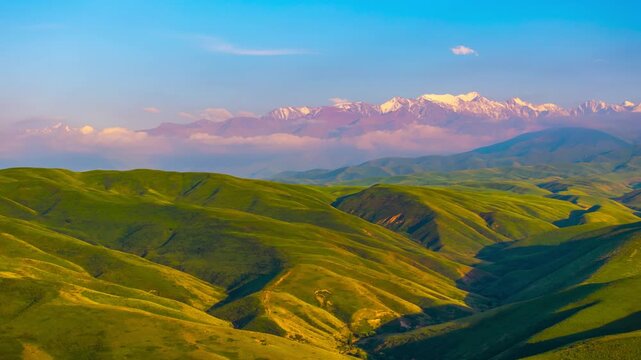 Drone hyperlapse flying forward above green foothills with snowcapped peaks on the horizon in Kyrgyzstan at summer evening, 70 mm telephoto angle of view.