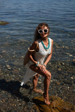 Summer lifestyle portrait of stylish woman wearing crochet dress and sunglasses while standing in shallow water at rocky beach