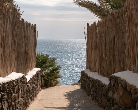 A narrow sandy path framed by rustic reed fences and stone walls leading to the sparkling blue sea on a bright, sunny day at a coastal resort