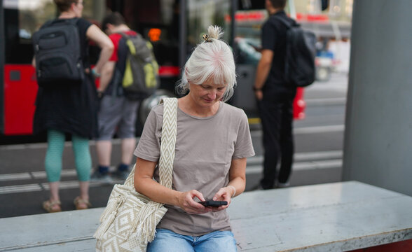 Senior woman with grey hair using smartphone while waiting at bus station in the city - Travel, elderly person lifestyle and technology concept