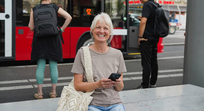 Happy senior woman with grey hair smiling on camera while using smartphone at bus station in the city - Travel, joyful elderly person and urban lifestyle concept
