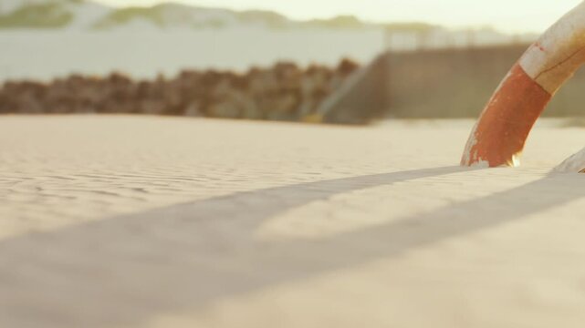 A lifeguard buoy is partially buried in the sand during low tide on the beach. Soft sunlight creates long shadows across the sand. The ocean can be seen in the background.