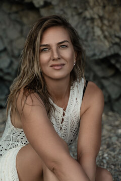 Summer fashion portrait of young woman wearing white lace dress sitting on rocky beach