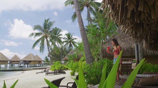 Tahiti hotel woman tourist drinking fruit drink cocktail at resort room balcony on beach. Vacation travel in Bora Bora, French Polynesia. Raw unedited video of real woman model without makeup