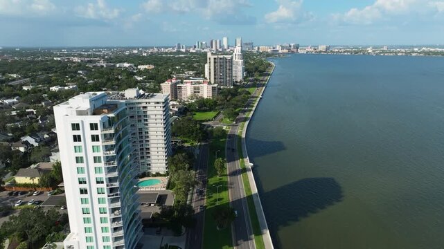 Drone perspective of the calm curve of Hillsborough Bay meeting Bayshore Boulevard in Tampa's Hyde Park, with waterfront condos and lush tropical foliage.