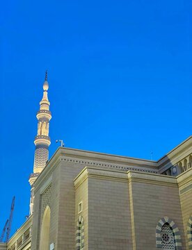 Low angle shot of an illuminated minaret and stone facade of Al-Masjid an-Nabawi against a vibrant blue sky.