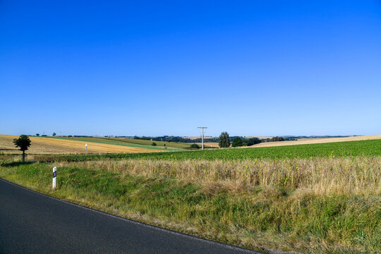 Landstra&szlig;e entlang weiter Felder unter intensiv blauem Himmel. L&auml;ndliche Sommerlandschaft mit Landwirtschaft, Roadtrip, Mobilit&auml;t, Natur, Weite und ruhiger Fahrt durch die Region.