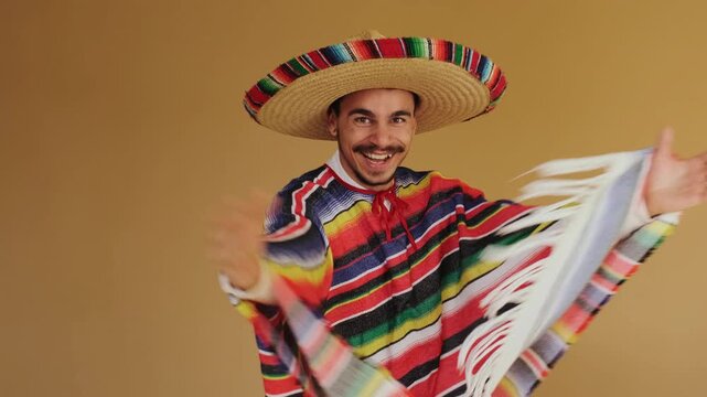 Young man wearing oversized straw sombrero with multicolored rim band and striped woven serape poncho spreads arms to display full garment wingspan while smiling and engaging camera against flat
