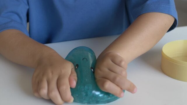 Child uses both hands to squeeze blue squishy toy on table, exploring its soft flexible texture. Sensory play, tactile stimulation, fine motor practice, hand coordination and focused activity.