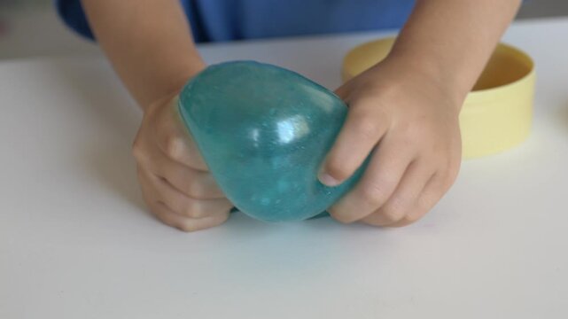 Young hands press and shape blue squishy toy during tabletop play. Soft texture, tactile exploration, sensory stimulation, hand coordination and calming repetitive movement create playful activity.