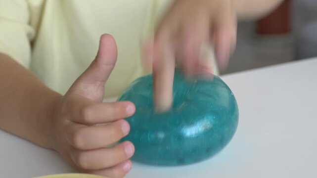Child presses glittery turquoise squishy character toy closeup at table during skill building with steady hand. Soft antistress toy supports motor development through playful squeeze activity