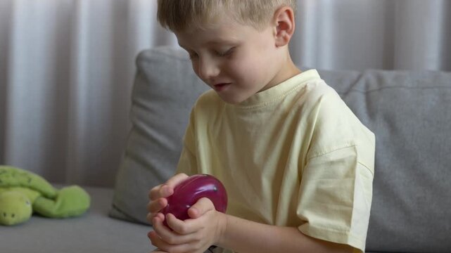 Young boy sits on sofa examining pink squishy toy with focused hands. Sensory play, tactile exploration, quiet concentration, fine motor practice and relaxing childhood activity indoors.