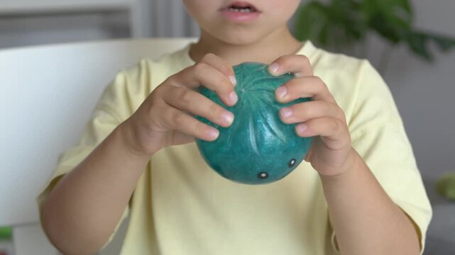 Boy holds sparkling teal squishy dumpling toy close to face showing joyful expression closeup in warm tone. Glitter texture and cute smiling character capture warm childhood emotion moment
