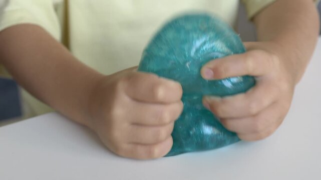 Child presses glittery turquoise squishy character toy closeup at table during skill building with steady hand. Soft antistress toy supports motor development through playful squeeze activity 